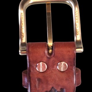 Peut inclure: Gros plan sur une ceinture en cuir marron avec une boucle dor&eacute;e. La boucle est rectangulaire avec un dessus arrondi. La ceinture comporte deux rivets argent&eacute;s et un motif estamp&eacute;. Les mots "MADE IN ITALY" et "SOLID BRASS" sont visibles.
