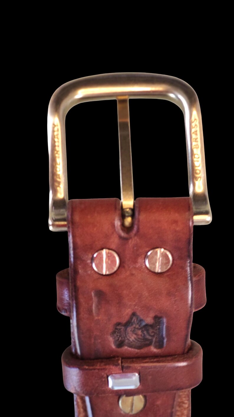 Peut inclure: Gros plan sur une ceinture en cuir marron avec une boucle dor&eacute;e. La boucle est rectangulaire avec un dessus arrondi. La ceinture comporte deux rivets argent&eacute;s et un motif estamp&eacute;. Les mots "MADE IN ITALY" et "SOLID BRASS" sont visibles.