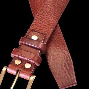 Peut inclure: Une ceinture en cuir marron avec une boucle dor&eacute;e. La ceinture a une surface textur&eacute;e et est fix&eacute;e avec des rivets de couleur laiton. La boucle est rectangulaire et la ceinture est pr&eacute;sent&eacute;e sur fond noir.
