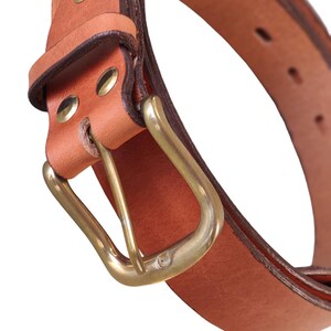 Puede incluir: Primer plano de un cintur&oacute;n de cuero marr&oacute;n con hebilla de lat&oacute;n. El cintur&oacute;n tiene un dise&ntilde;o cl&aacute;sico con hebilla rectangular y est&aacute; asegurado con remaches de lat&oacute;n. El cuero tiene un aspecto natural y texturizado.
