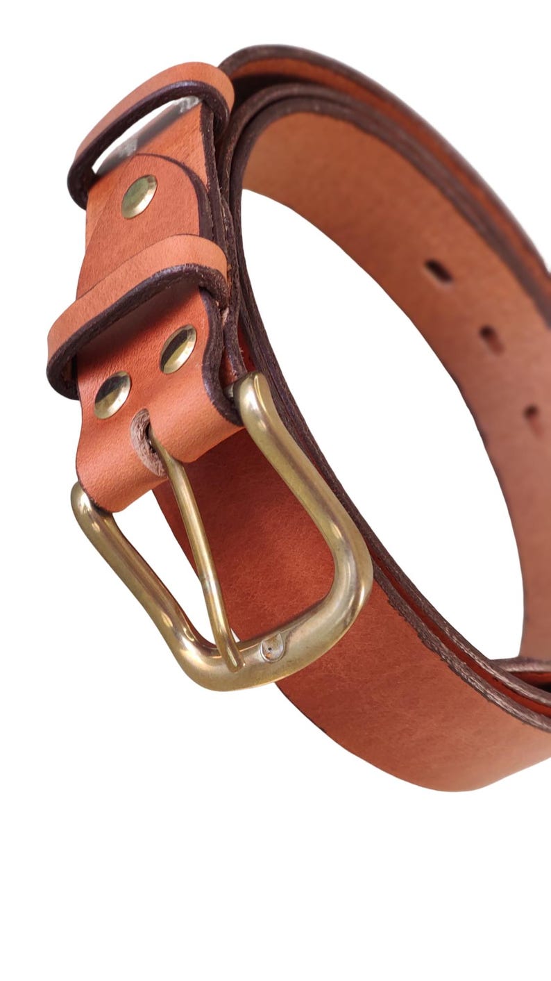 Puede incluir: Primer plano de un cintur&oacute;n de cuero marr&oacute;n con hebilla de lat&oacute;n. El cintur&oacute;n tiene un dise&ntilde;o cl&aacute;sico con hebilla rectangular y est&aacute; asegurado con remaches de lat&oacute;n. El cuero tiene un aspecto natural y texturizado.