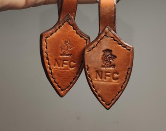 Puede incluir: Dos llaveros de cuero marrón con anillas y cierres plateados. Cada llavero tiene una etiqueta en forma de escudo con un logotipo y el texto "NFC".