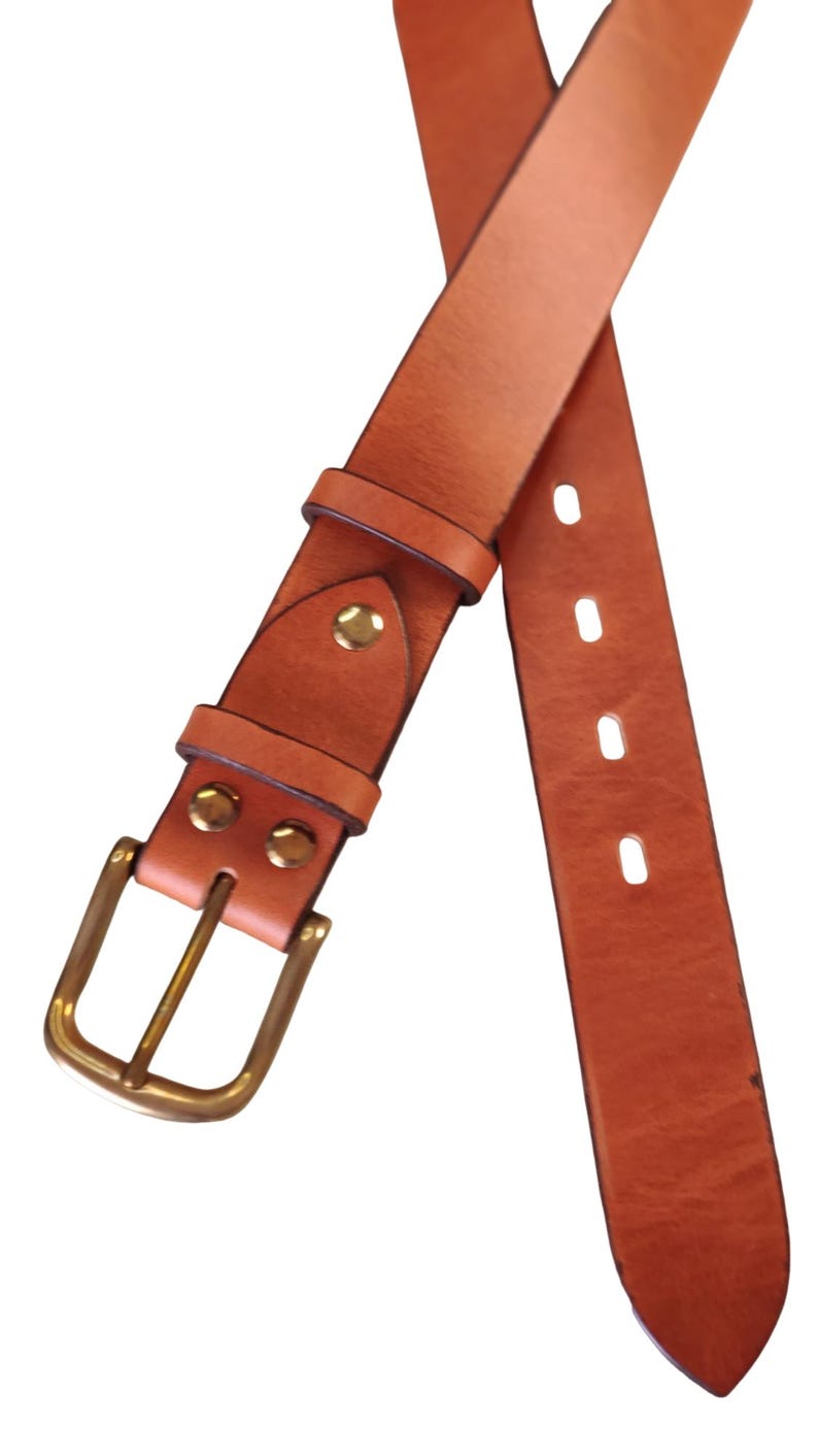 Puede incluir: Un cintur&oacute;n de cuero marr&oacute;n claro con hebilla y remaches de lat&oacute;n. El cintur&oacute;n tiene un dise&ntilde;o cl&aacute;sico con una hebilla rectangular y m&uacute;ltiples agujeros para ajustar. El cuero parece ser de alta calidad, con una textura suave y un color c&aacute;lido y natural.