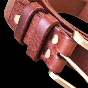 Op de afbeelding: Close-up van een bruine leren riem met een messing gesp en klinknagels. De riem is gemaakt van dik, getextureerd leer en heeft een klassiek ontwerp. De messing gesp voegt een vleugje elegantie toe.