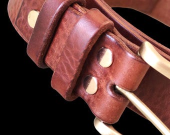 Puede incluir: Primer plano de un cinturón de cuero marrón con hebilla y remaches de latón. El cinturón está hecho de cuero grueso y texturizado y tiene un diseño clásico. La hebilla de latón añade un toque de elegancia.