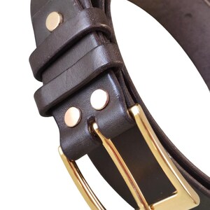 K&ouml;nnte beinhalten: Nahaufnahme eines braunen Lederg&uuml;rtels mit goldfarbener Schnalle. Der G&uuml;rtel hat zwei goldene Nieten in der N&auml;he der Schnalle und eine Schlaufe, um den G&uuml;rtel an Ort und Stelle zu halten. Der G&uuml;rtel ist gebogen und scheint neu zu sein.