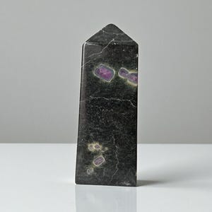 Può includere: Un obelisco verde scuro con inclusioni viola e gialle. La pietra ha una forma rettangolare con la parte superiore appuntita. La superficie presenta crepe visibili e una finitura lucida. Questo oggetto decorativo è adatto all'arredamento della casa.