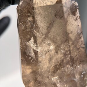 May include: A large, translucent smoky quartz crystal with a faceted, pointed tip. The crystal has a light brown color with internal inclusions and a slightly rough texture. The image is a close-up, highlighting the natural beauty and clarity of the gemstone.