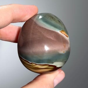 May include: A polished, oval-shaped stone with a smooth surface. The stone features bands of color, including shades of brown, green, and white. The stone is held between two fingers.