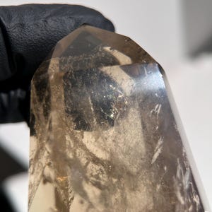 May include: A translucent, smoky quartz crystal with a polished, faceted point. The crystal has a light brown color and appears to be held by a black gloved hand. The crystal's surface has internal fractures and inclusions.