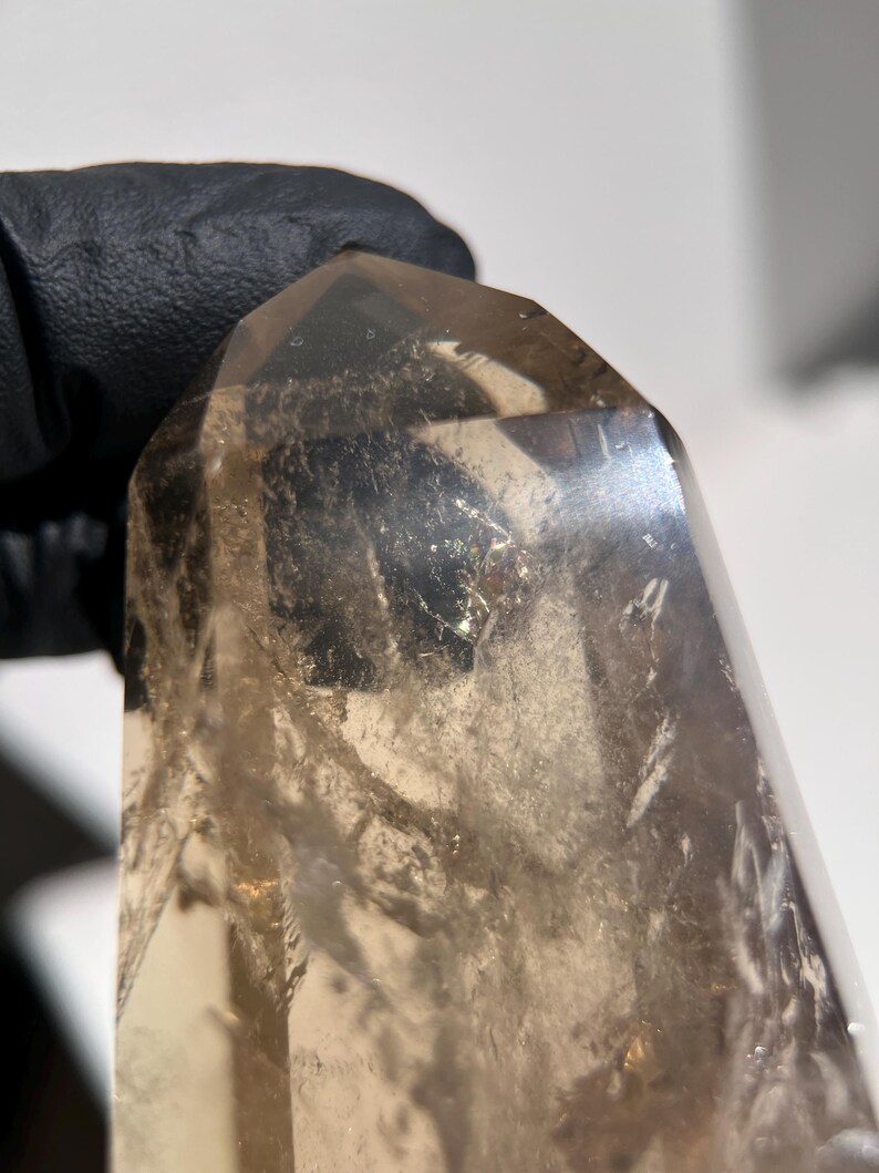 May include: A translucent, smoky quartz crystal with a polished, faceted point. The crystal has a light brown color and appears to be held by a black gloved hand. The crystal's surface has internal fractures and inclusions.