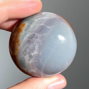 Op de afbeelding: Een gepolijste edelsteenbol met een glad, reflecterend oppervlak. De bol heeft een mix van kleuren, waaronder grijstinten, bruin en wit. De steen wordt tussen twee vingers gehouden, wat de grootte en vorm laat zien.