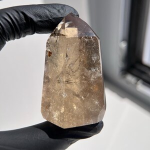 May include: A faceted, smoky quartz crystal point. The crystal is held by a gloved hand. The crystal is a translucent brown color with internal inclusions and a polished surface. The crystal has a hexagonal base and tapers to a point.