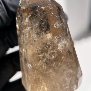 May include: A faceted, smoky quartz crystal point. The crystal is a translucent brown color with internal inclusions and a polished surface. The crystal is held against a white background.