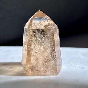 May include: A clear, faceted crystal point with a light brown hue. The crystal has a polished, smooth surface and a pointed top. The crystal is standing upright on a white surface, with a dark background.