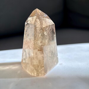 May include: A faceted, light brown crystal point. The crystal is a translucent amber color, with visible internal structures and a polished, geometric shape. The crystal is standing upright on a white surface.