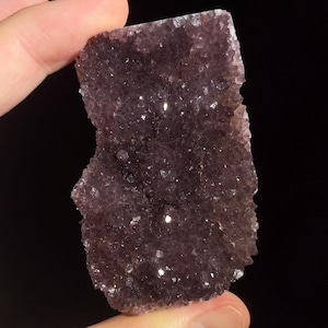 May include: A dark purple amethyst crystal cluster with a rough, textured surface. The crystal has a natural, irregular shape and is held against a black background. The surface sparkles with small, reflective facets.