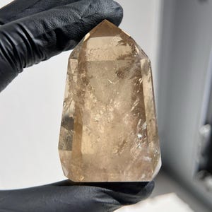 May include: A polished, faceted, smoky quartz crystal point. The crystal is a translucent, light brown color with internal inclusions. The point is held by a gloved hand against a white background. The crystal is a natural, geological specimen.