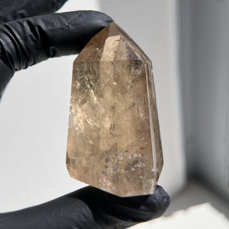 May include: A faceted, light brown crystal tower is held in a black gloved hand. The crystal has a rectangular base and tapers to a pointed top. The crystal is translucent with internal inclusions.