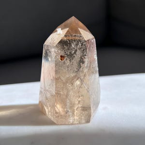 May include: A faceted, light brown crystal point with a polished finish. The crystal has a tapered shape and a pointed top. The crystal is translucent, allowing light to pass through. The crystal is a natural mineral specimen.