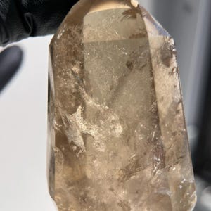 May include: A translucent, light brown crystal point with a faceted shape. The crystal has internal inclusions and a polished surface. The crystal is held by a gloved hand. The crystal is a natural mineral specimen.