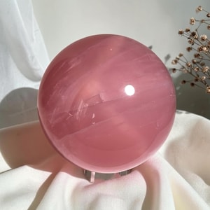 Op de afbeelding: Een gepolijste, bolvormige rozenkwarts kristal met een glad, doorschijnend oppervlak. De steen vertoont een zachte roze tint met subtiele interne aders en natuurlijke insluitingen, die het licht reflecteert met een heldere, ronde glans.