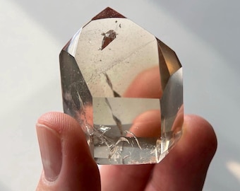 May include: A clear quartz crystal with a brown tip and inclusions. The crystal is faceted and held in a hand, showcasing its natural beauty. The crystal is a light brown color and has a geometric shape.