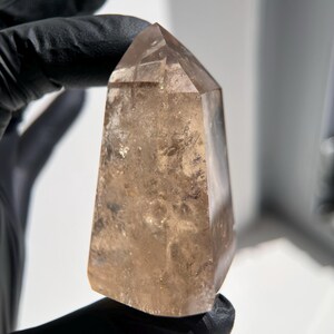 May include: A polished, light brown quartz crystal point. The crystal has a tapered shape with a flat, faceted top. The surface of the crystal is translucent, with subtle variations in color and texture. The crystal is held by a gloved hand.