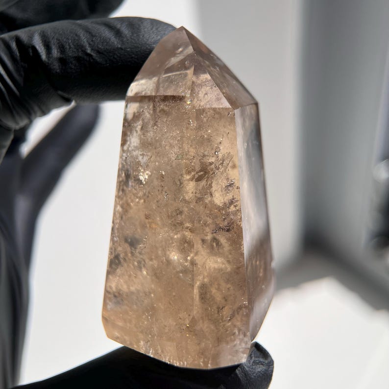 May include: A polished, light brown quartz crystal point. The crystal has a tapered shape with a flat, faceted top. The surface of the crystal is translucent, with subtle variations in color and texture. The crystal is held by a gloved hand.