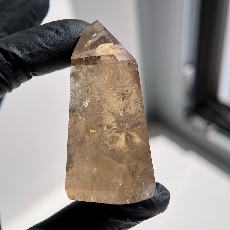 May include: A polished, light brown quartz crystal point. The crystal has a smooth, faceted surface and a tapered shape, with a pointed top. The crystal is held by a gloved hand, and the background is blurred.