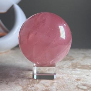 Può includere: Una sfera rosa traslucida e lucidata in quarzo rosa, che mostra fratture interne naturali e variazioni di colore. La sfera è montata su un piccolo supporto quadrato trasparente in vetro o acrilico, che fornisce una base stabile per l'esposizione.