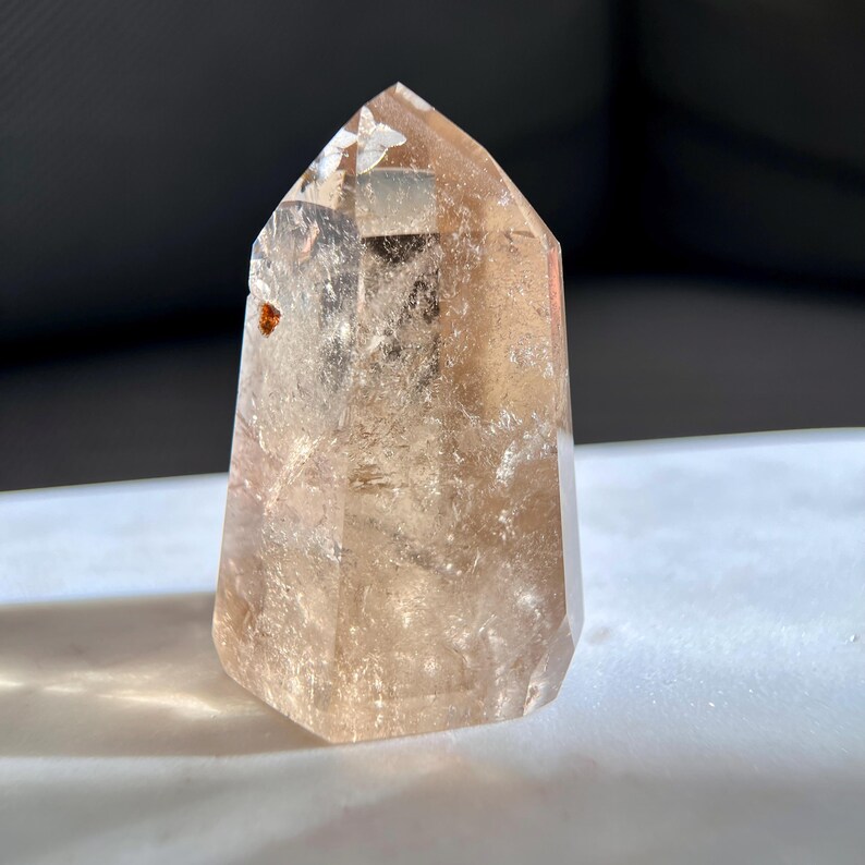 May include: A clear, faceted crystal point with a light brown hue. The crystal has a hexagonal base and tapers to a pointed top. The surface has internal inclusions and imperfections. The crystal is displayed on a white surface.