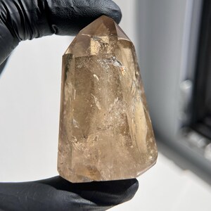 May include: A clear, brown quartz crystal point. The crystal is held by a gloved hand. The crystal has a faceted top and a smooth, polished surface. The color is a light, translucent brown with some internal inclusions.