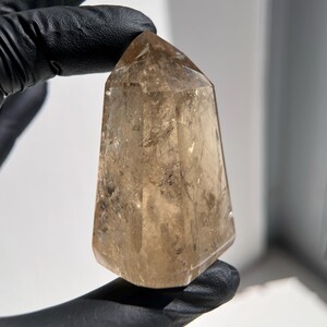 May include: A polished, light brown quartz crystal point. The crystal is held by a gloved hand. The crystal has a smooth, faceted surface and a tapered shape, with a pointed top. The crystal is translucent, allowing light to pass through.
