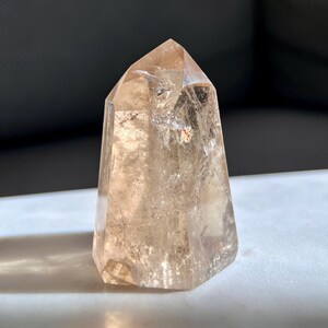 May include: A clear, faceted crystal point with a light brown hue. The crystal has a hexagonal base and tapers to a sharp point. The surface has internal fractures and inclusions, catching the light. This is a decorative crystal for display.