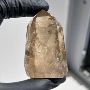May include: A faceted, amber-colored crystal point. The crystal is held by a gloved hand, showcasing its natural, translucent beauty. The crystal's surface has subtle variations in color and texture, reflecting light.