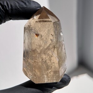 May include: A clear, faceted smoky quartz crystal point. The crystal is a light brown color with a pointed top and a small, golden inclusion. The crystal is held by a gloved hand.