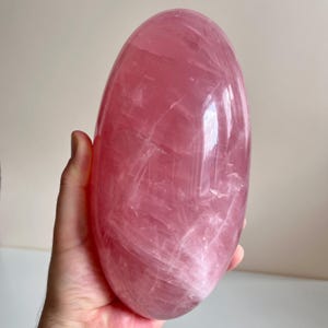 Könnte beinhalten: Ein großer, glatter, oval geformter Rosenquarzkristall mit polierter Oberfläche. Der Stein zeigt einen durchscheinenden, blassrosa Farbton mit sichtbaren inneren Brüchen und natürlichen Adern in seiner Struktur, die das Licht reflektieren.