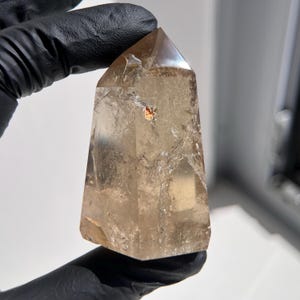 May include: A clear, light brown quartz crystal point. The crystal is held by a gloved hand. The crystal has a faceted top and a slightly rough base. The crystal has a few inclusions and internal fractures.