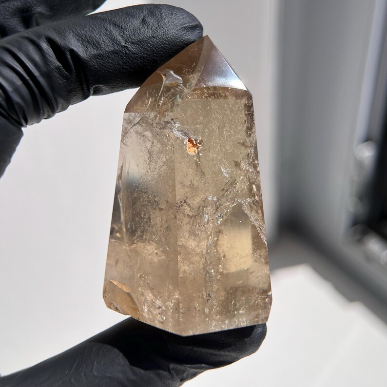 May include: A clear, light brown quartz crystal point. The crystal is held by a gloved hand. The crystal has a faceted top and a slightly rough base. The crystal has a few inclusions and internal fractures.