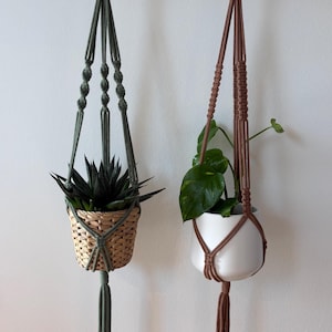Könnte beinhalten: Zwei Makramee-Pflanzenaufhänger. Einer ist olivgrün und hält einen geflochtenen Korb mit einer Sukkulente. Der andere ist braun und hält einen weißen Keramiktopf mit einer grünen Blätterpflanze. Beide sind an einem Holzbalken aufgehängt.