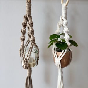 Könnte beinhalten: Zwei Makramee-Pflanzenaufhänger. Einer ist beige und hält ein klares Glas mit einer Kerze. Der andere ist weiß und hält einen kleinen braunen Topf mit einer grünen Pflanze. Beide haben Holzringe zum Aufhängen.