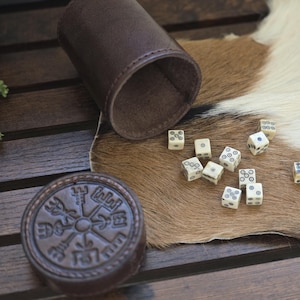May include: A dark brown cylindrical leather dice cup with a matching circular lid embossed with a compass-like symbol. Ten small, off-white cubic dice with black circular markings are scattered nearby, featuring various patterns of dots and rings.