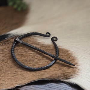 Puede incluir: Una broche de anillo de metal negro con un dise&ntilde;o de alambre retorcido y un pasador largo y recto. La broche est&aacute; sobre una superficie de piel marr&oacute;n y blanca.