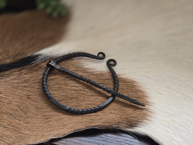 Puede incluir: Una broche de anillo de metal negro con un dise&ntilde;o de alambre retorcido y un pasador largo y recto. La broche est&aacute; sobre una superficie de piel marr&oacute;n y blanca.