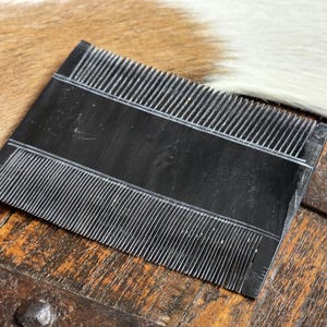 May include: A black metal comb with fine teeth, designed for grooming animal fur. The comb is lying on a wooden surface.