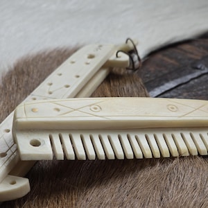 May include: A white bone comb with a decorative handle. The comb is folded in half and has a carved design on the handle. The comb is lying on a brown fur surface.