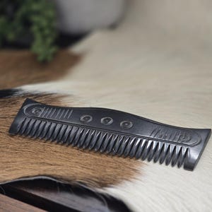 Puede incluir: Un peine gris oscuro con un mango curvo y dientes finos. El peine presenta grabados circulares y lineales decorativos. Se muestra sobre una alfombra de piel tricolor.