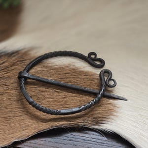 Puede incluir: Un broche de estilo vikingo de metal negro con un dise&ntilde;o de alambre retorcido y un bucle decorativo en el extremo. El broche est&aacute; sobre una superficie de piel marr&oacute;n y blanca.