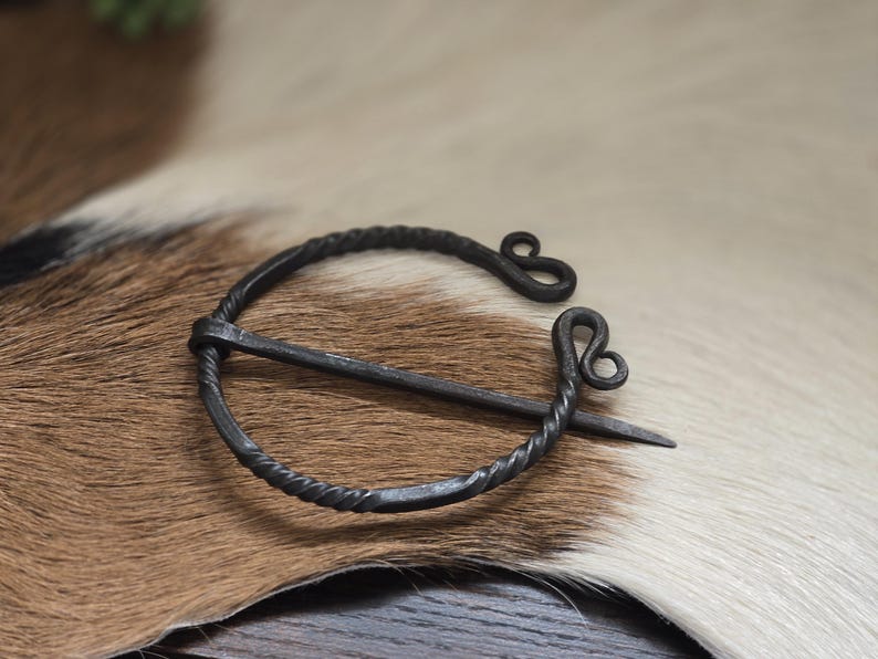 Puede incluir: Un broche de estilo vikingo de metal negro con un dise&ntilde;o de alambre retorcido y un bucle decorativo en el extremo. El broche est&aacute; sobre una superficie de piel marr&oacute;n y blanca.